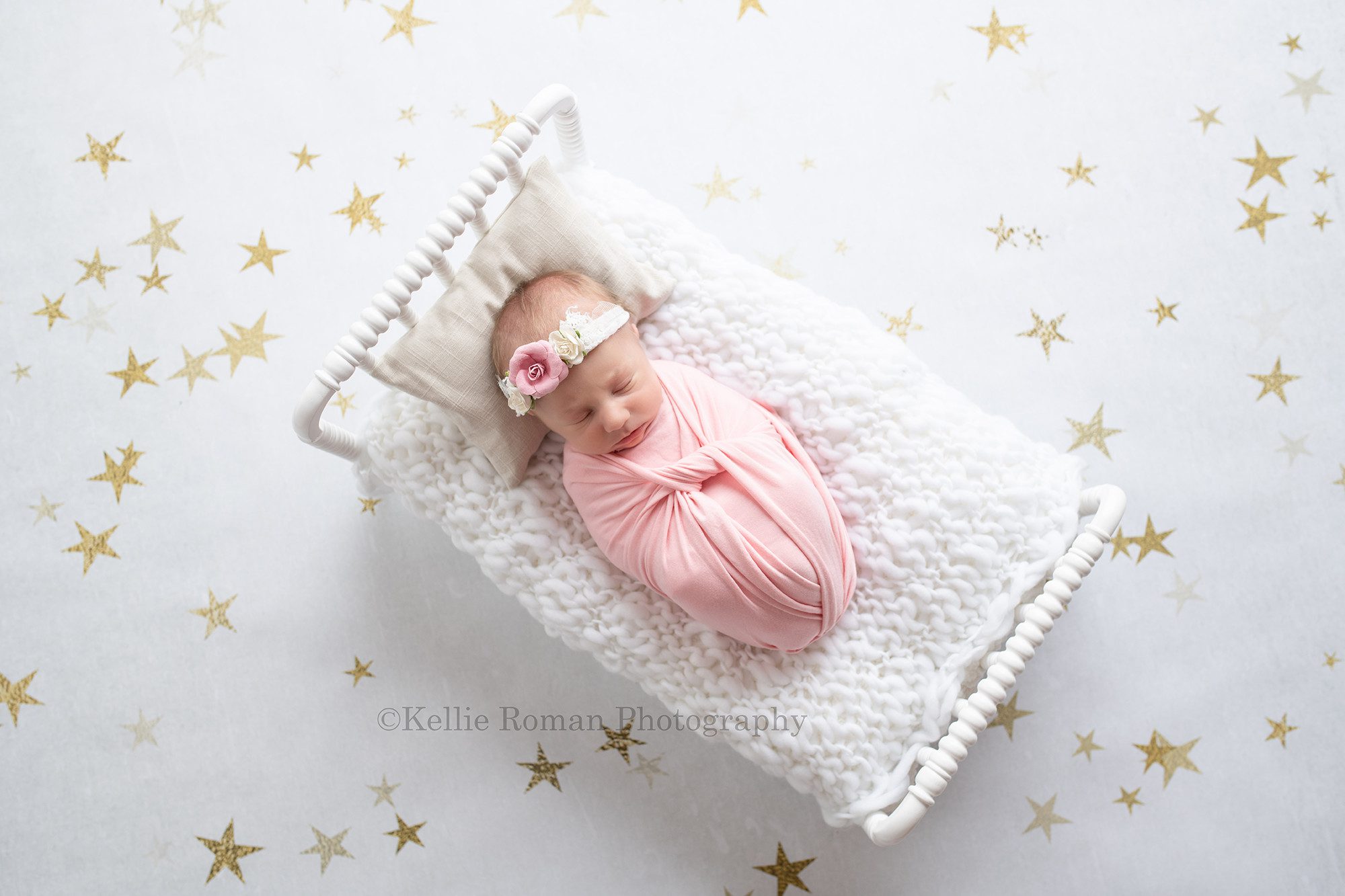 milwaukee baby whisperer a newborn baby girl is in a greendale Wisconsin photo studio. She is wrapped in a pink fabric and has a pink and white flower headband on. she is laying on a white little bed with white knitted fabric on it. her head is resting on a pillow. the best is on top of a white backdrop with gold stars.