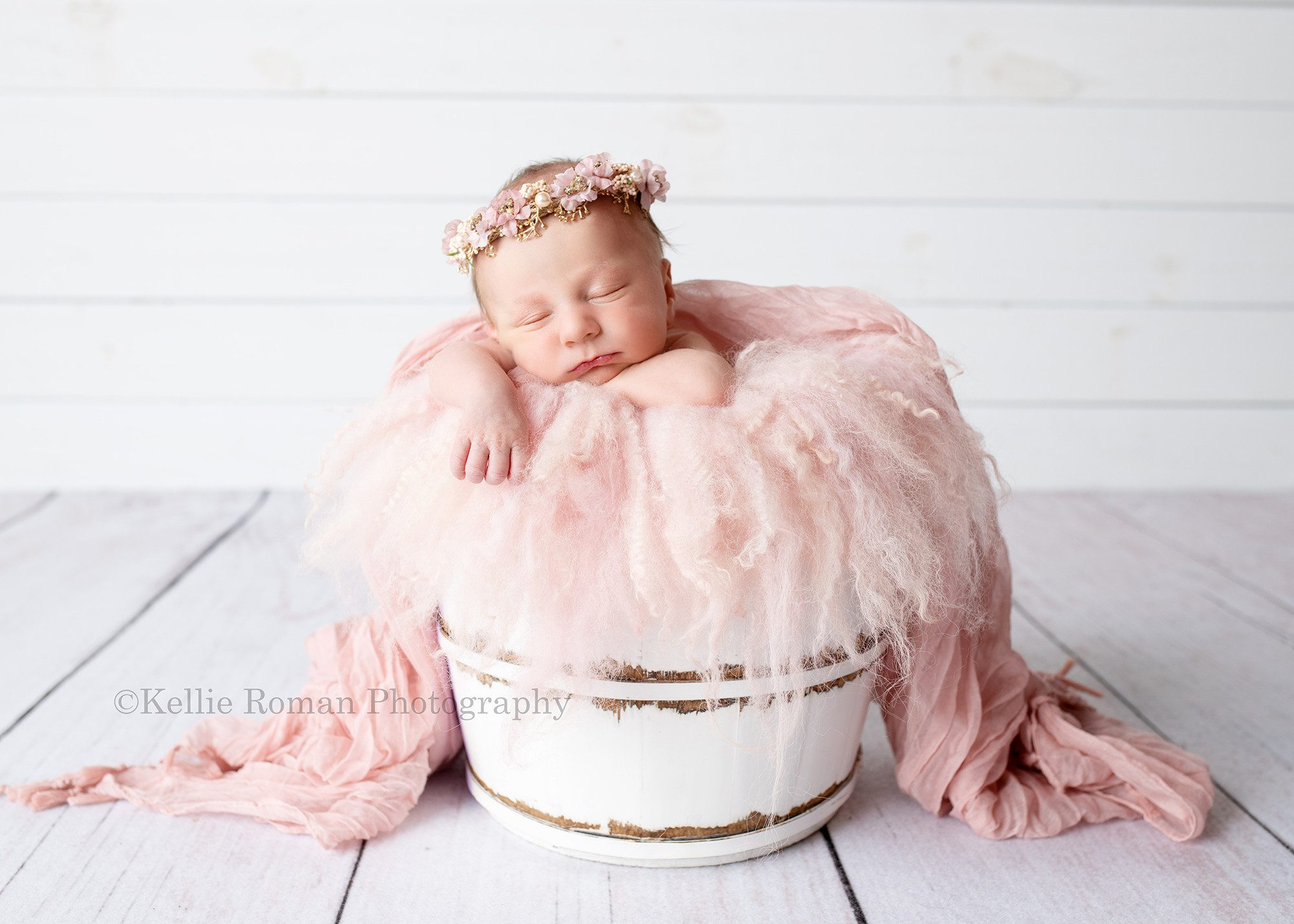 milwaukee baby whisperer a newborn baby girl in a milwaukee photogorapher studio she is posed upright in a white wood bucket filled with light pink fur and fabric. She has one arm hanging over the edge of the bucket and the other is under her chin she has a pink and gold headband on and is in front of a white wood wall with white wood floor under her.