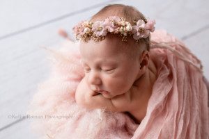 milwaukee baby whisperer a newborn baby girl is being photographed in a Milwaukee Wisconsin photographers studio. she is in a bucket filled white light pink fluff fur. She has her chin resting on her arms and is wearing a pink and gold floral headband.