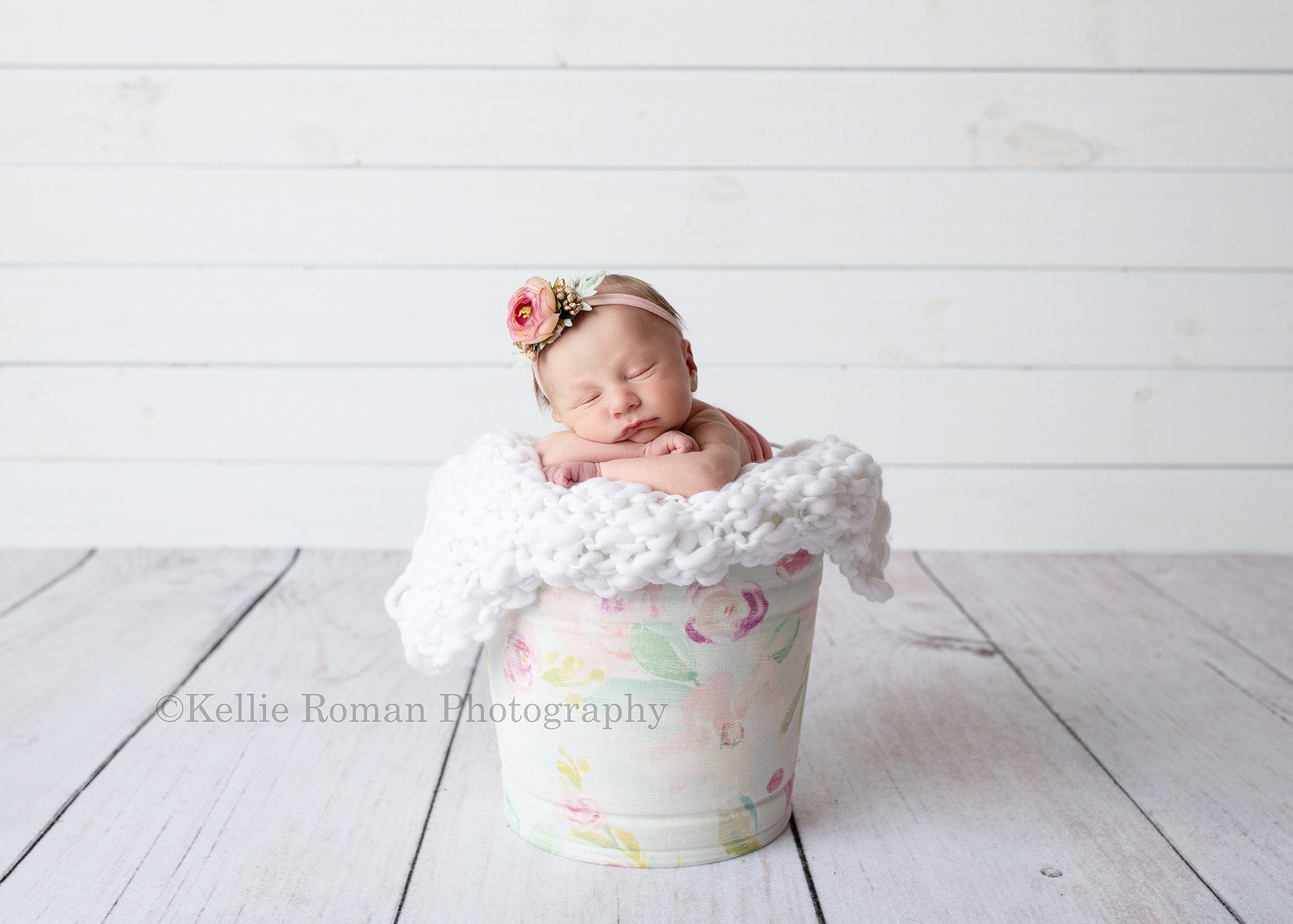 milwaukee baby whisperer a newborn infant girl in a milwaukee photographers studio she is posed in a vintage floral bucket with a white knitted blanket in it. she has her chin resting on her arms in the bucket and she has a pink and gold flower headband on. the floor is white wood, and she is in front of a white wood shiplap backdrop.