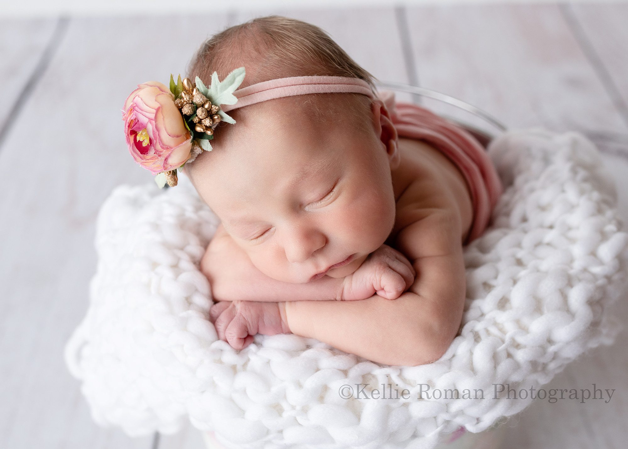 milwaukee baby whisperer a sweet baby girl in a milwaukee photographers studio is sleeping while posed upright in a bucket. She has her chin resting on her hands and is on a white knitted blanket. She has on a pink and gold flower headband.