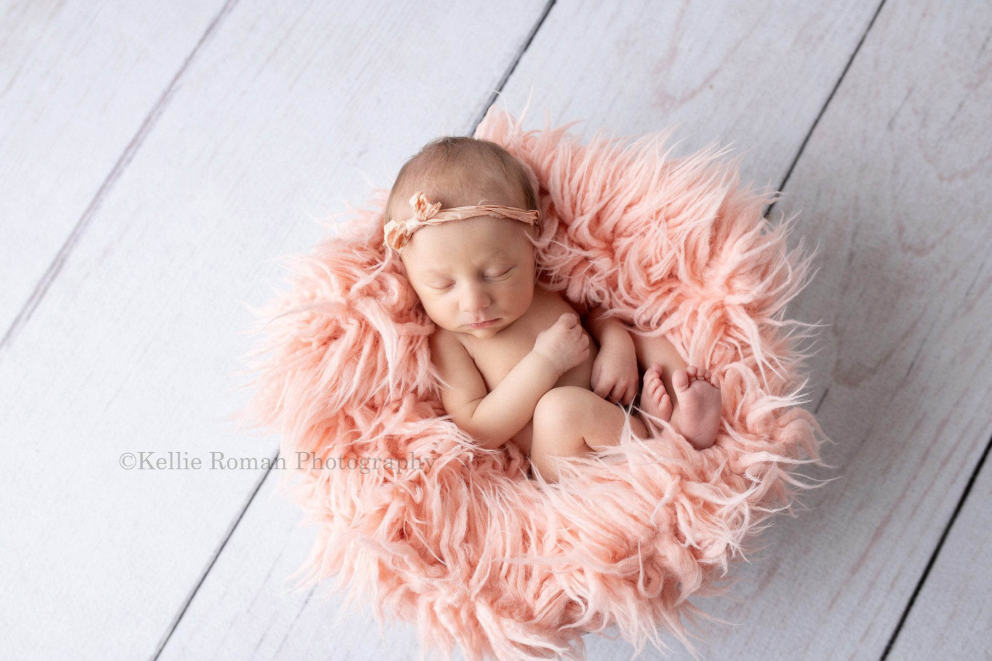 milwaukee baby whisperer a newborn baby girl in a milwaukee photographers studio is naked in a wood tub with pink fur. The newborn is curled up and sleeping and she has on a pink bow headband. She's on top of white wood floor.