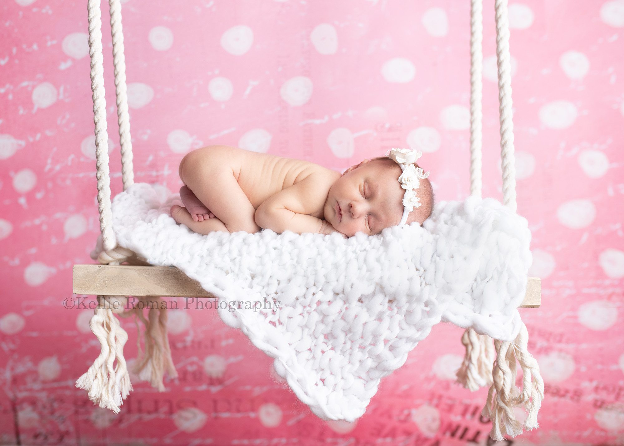 milwaukee baby whisperer a newborn baby girl in a milwaukee photo studio she is laying on her belly naked on a wood swing the swing has a white fabric on it and it's hanging in front of a pink and white polka dot backdrop