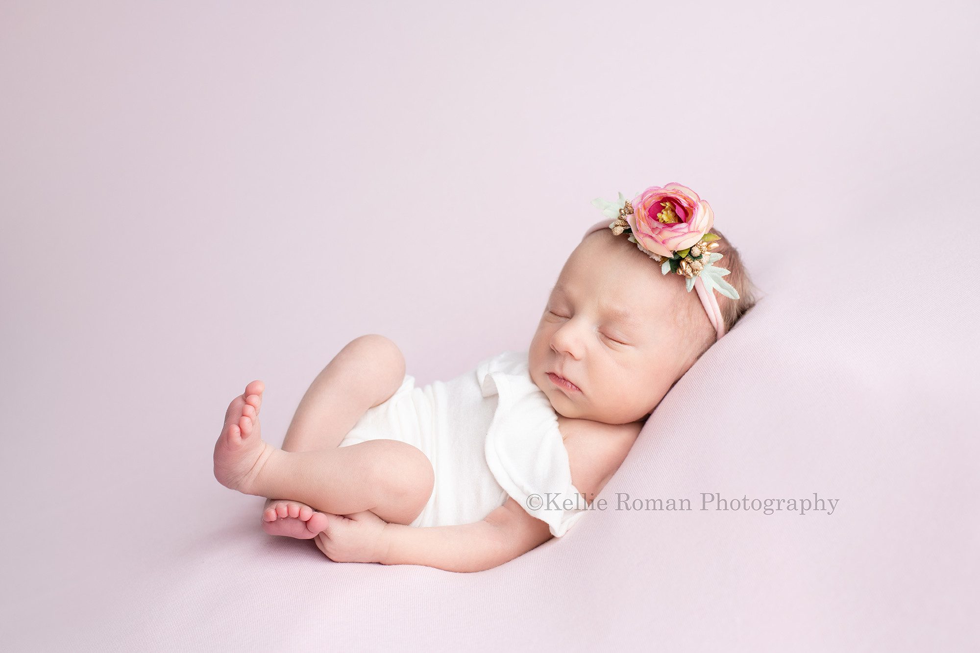 milwaukee baby whisperer a newborn baby girl in a greendale photographers studio she is sleeping while laying on her back wearing a white romper and a purple flower headband. She's laying on a pink fabric and is very relaxed.