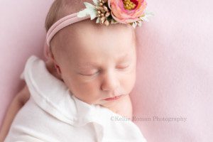 milwaukee baby whisperer a close up shot of a newborn baby girl in a milwaukee photographers studio she has a white romper on and a purple flower headband. She's sleeping on top of a pink blanket.