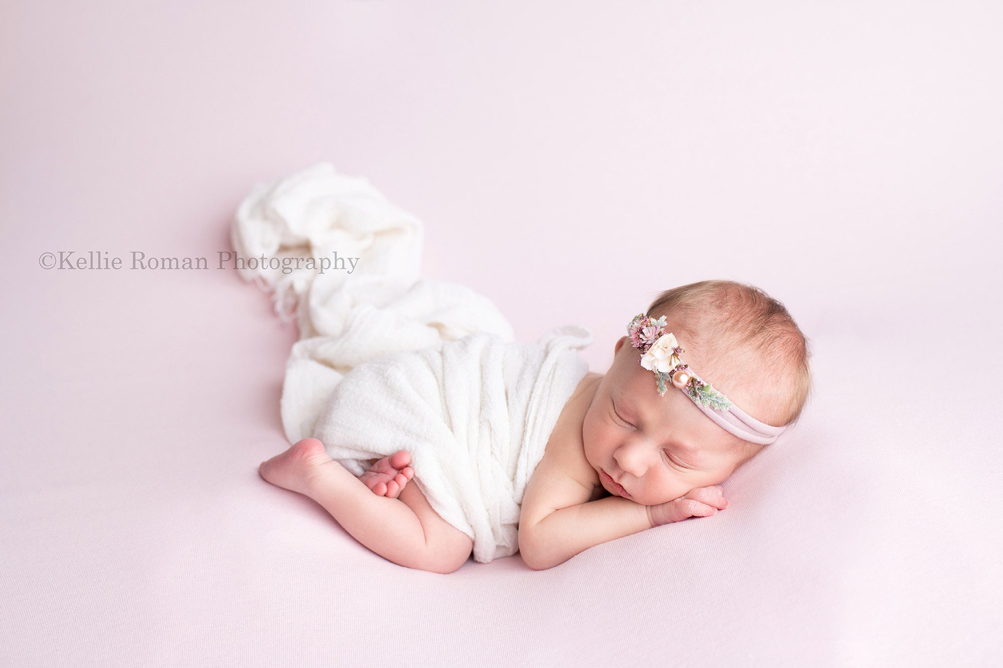 milwaukee newborn whisperer a newborn baby girl in a milwaukee photography studio she is sleeping on her tummy on top of a pink posing blanket she has a white wrap covering her diaper and back and a light purple flower headband on