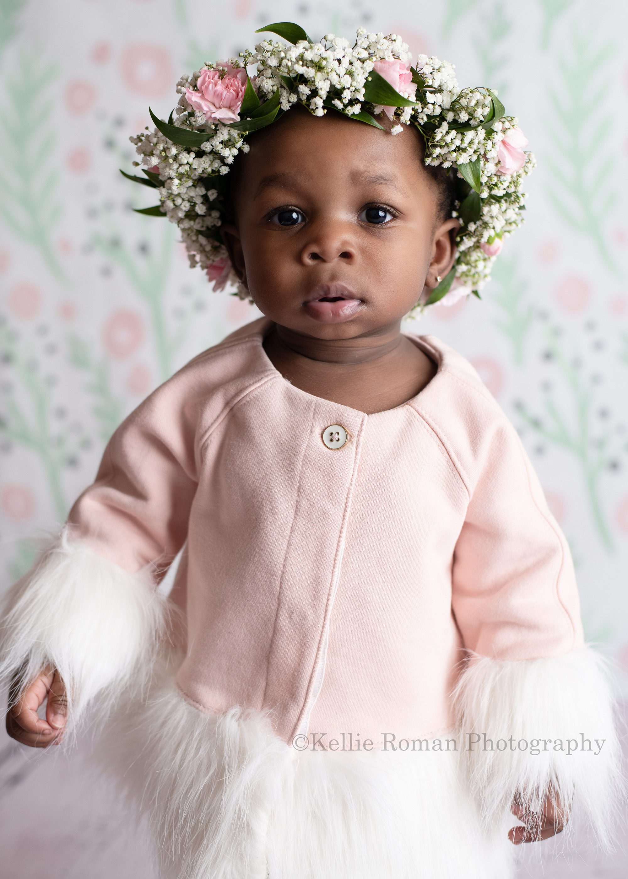 takes the cake. a one year old girl in Milwaukee photographers studio in Wisconsin is looking into the. camera. it's a close up shot of her face and she is wearing a light pink sweater with white fur along the sleeves. She has a floral crown on.