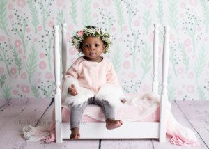 takes the cake a one year old little girl in a milwaukee photographers studio she is sitting on a white four post bed that has white and light pink fabric covering the mattress the bed is on a white wood floor in front of a pastel pink grey and green floral backdrop the girl is wearing a light pink sweater with white fur on the sleeves along with grey pants. she has a real flower headband on and is looking at the camera