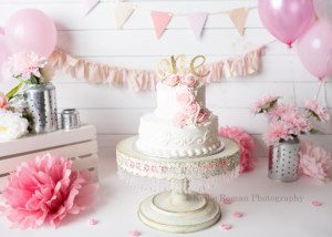 takes the cake a shot of a one year old cake and cake smash set up the cake is white and two tiers with light pink flowers. the cake is on a huge ivory cake stand with crystals on the bottom. the entire scene has light pink dark pink and white flowers in metal lanterns, pink and ivory backdrops and pink tissue paper pom poms