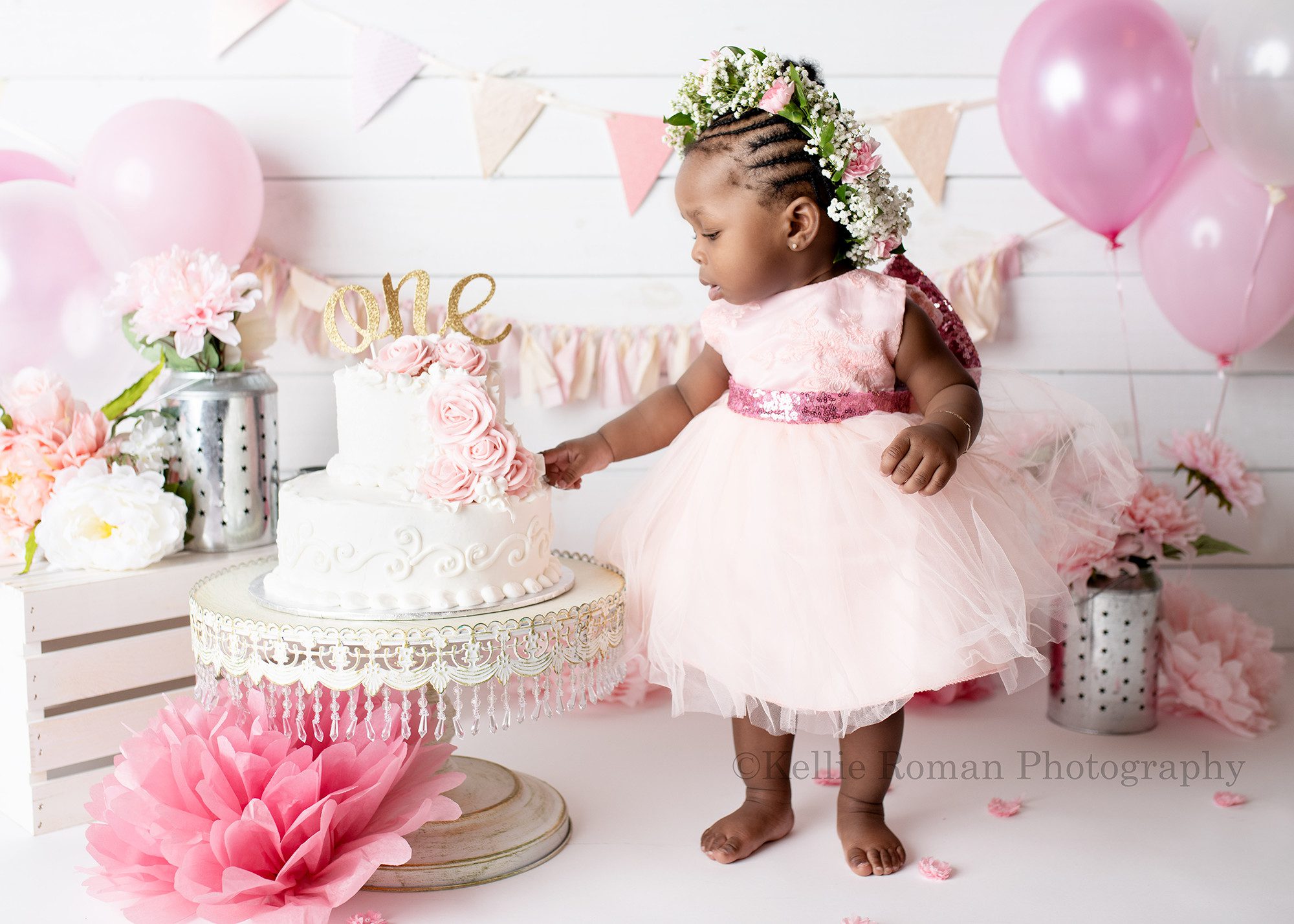 takes the cake a one year old girl is in a milwaukee photographers studio for her cake smash session she has a two tier white cake with pink flowers on a white cake stand with jewels she has to stand next to it it's so large. the girl has a pink dress on and the entire scene is filled with pink flowers and metal lanterns.