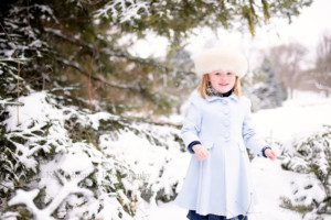 milwaukee snowfall a 5 year old girl is standing among huge pine trees covered in a fresh snow fall. She's wearing a baby blue pea coat with big buttons and scallops along the edges. She has a big white fur hat on and is looking at the camera a smiling. She has blonde hair and blue eyes.