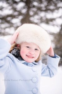 milwaukee snowfall. A 5 year old girl in a Milwaukee County park is surrounded by snow and pine trees. She's wearing a powder blue pea coat with scallops and a white fur hat. She's looking and smiling at the camera while holding onto her white fur hat. She has blonde hair and blue eyes