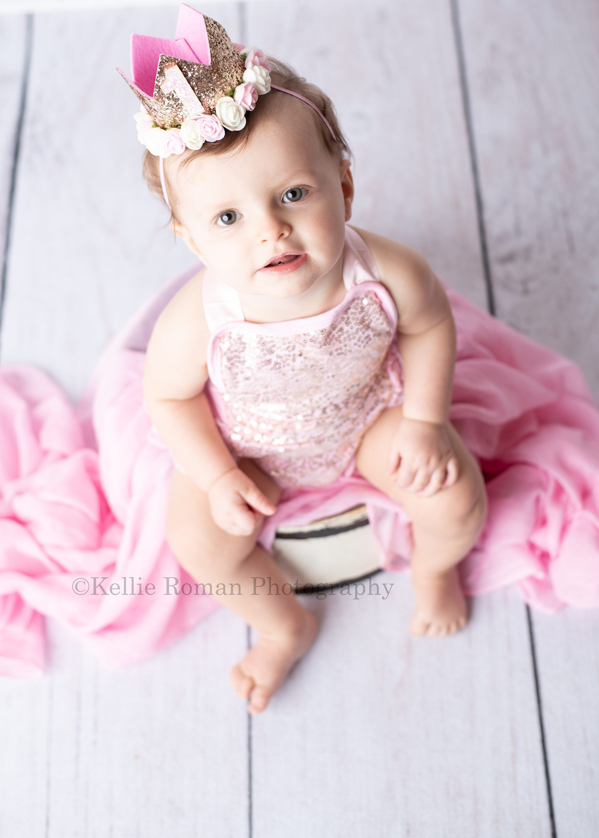 girlie cake smash a one year old girl is in a milwaukee photographers studio she's wearing a pink romper with gold sequins on the front. She's sitting on a white wood tub, and is looking up and smiling at the camera. the shot is very bright and white and airy.