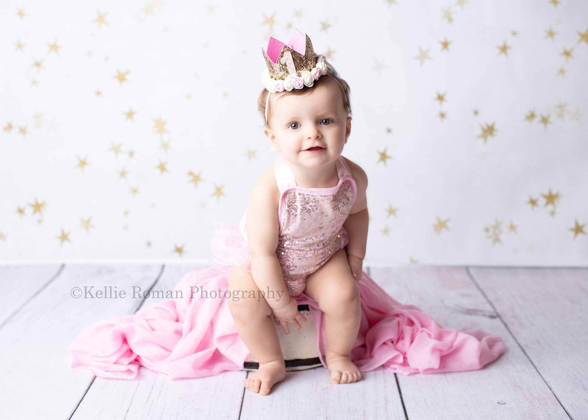 girlie cake smash a one year old girl is in a milwaukee photographers s studio she is sitting on a white wood tub with a pink fabric over it. She has a pink romper on with gold sequins and a gold crown hat. she's smiling at the camera. The floor is white wood and the backdrop is white with gold stars.