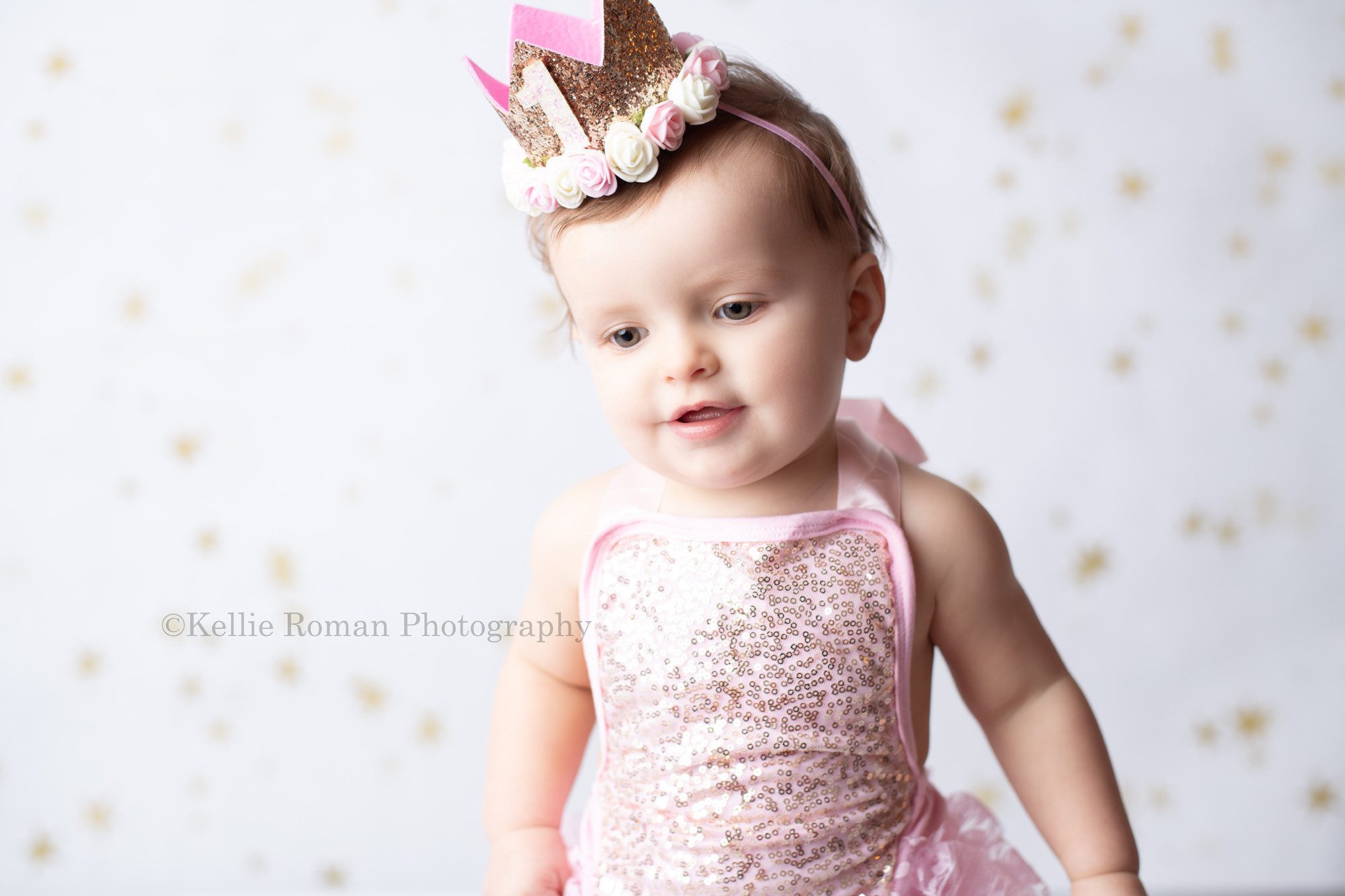 girlie cake smash a one year old girl is in a milwaukee photographers studio she is waring a pink romper with gold sequins and a gold crown headband. She is standing up but looking at the grown in front of a white backdrop with gold stars.