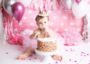 girlie cake smash a one year old girl in a milwaukee photographers studio smashing into a sprinkle cake the backdrop is very festive with bright pinks polka dots, pearls, and white lace banner. The one year old girl has frosting all over her face.