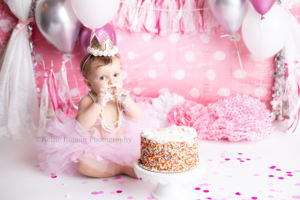 girlie cake smash a one year old little girl is in a milwaukee photographers studio. She is smashing into a white frosted cake with sprinkles. She's wearing a pink tutu with white pearl necklace and a crown headband. She has frosting all over her hands and face and is looking into the camera. The backdrop is very festive birthday with a pink polka dot backdrop, lace and pink banner and silver white and pink balloons.