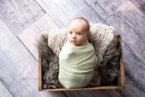 studio newborn session a infant baby boy is in a milwaukee photographers studio. He is wrapped in a lime green fabric, and is laying on his back in a wood crate filled with tan fabric and brown fur. He is wide awake and looking at the camera. He's on top of a dark wood floor.