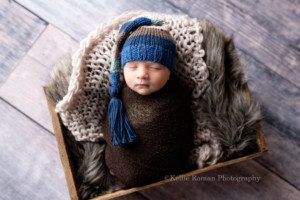 studio newborn session a infant boy is sleeping white laying on his back and swaddled in a brown fabric. The baby is inside of a wood crate filled with knit fabrics and a brown fur. The baby is wearing a blue tan and brown striped hat