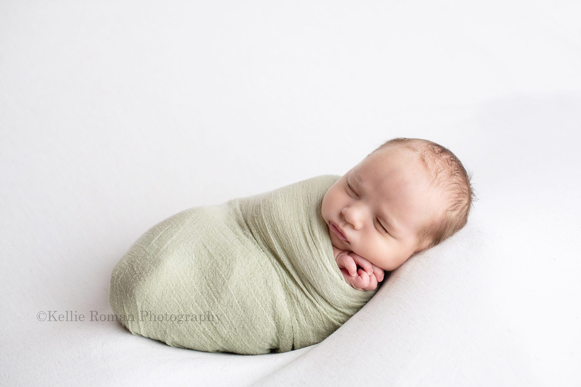 studio newborn session a baby boy is in a milwaukee photo studio located in greendale Wisconsin. The boy is laying on top of a white fabric and is wrapped in a green swaddle. His hands are poking up through the top by his chin and he is sleeping.