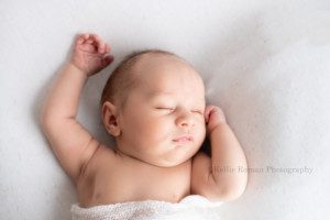 studio newborn session a baby boy is laying on his back on top of a cream colored blanket. he has a white swaddle wrap around his waist and below his arms are above his head and he is sleeping while his head is turned to one side