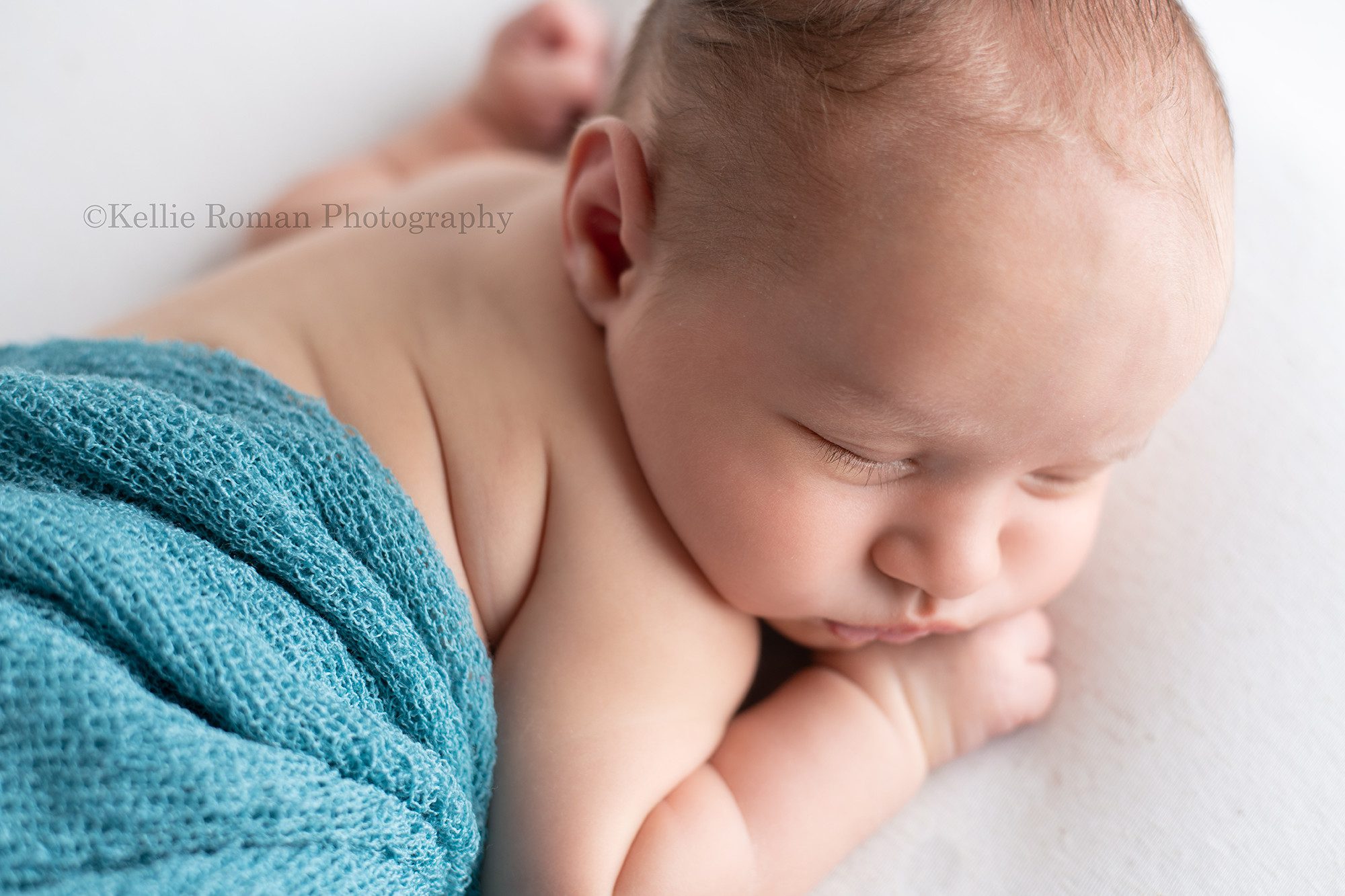 studio newborn session a baby. boy in a milwaukee photographers studio is sleeping on a bean bag covered with white fabric he has a teal wrap cover his diaper and the shot is taken from above his head