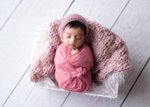 all wrapped up. A newborn baby girl in a pink swaddle and a pink mohair bonnet is asleep in a Milwaukee Wisconsin photographers studio. she is laying in a white wire basket filled with white fur and a pink knit blanket