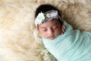 all wrapped up. a baby girl is in a Milwaukee Wisconsin photographers studio. the studio is in greendale Wisconsin. the baby is wrapped tight in a teal fabric and she has a few fingers sticking out of the top. she has a teal and white flower headband on, and lots of dark thick hair. the baby is resting on top of a cream colored rug