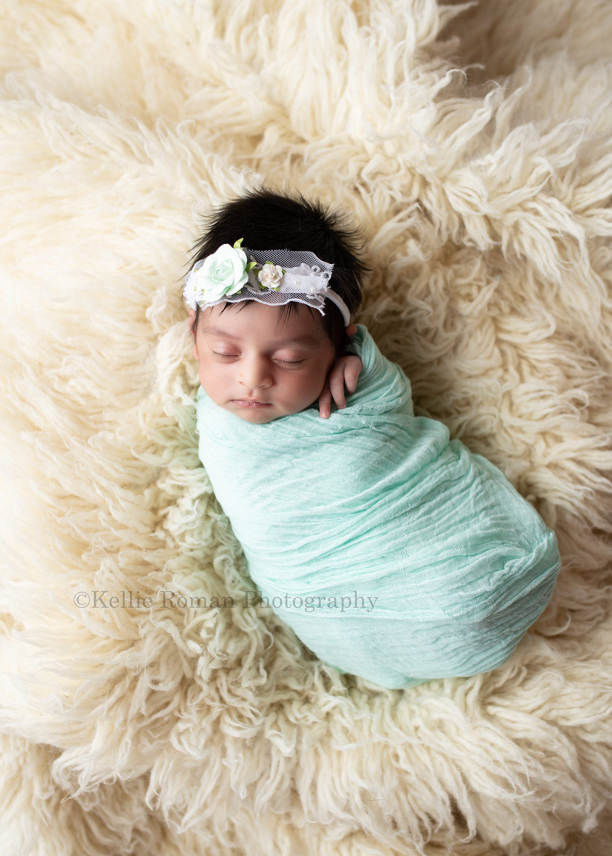 all wrapped up a newborn little baby girl is in a milwaukee photographer studio. the studio is in greendale Wisconsin. the baby is wrapped tight in a teal swaddle fabric and has on a white and teal flower headband. the baby girl is sleeping onto of a cream color flakoti rug