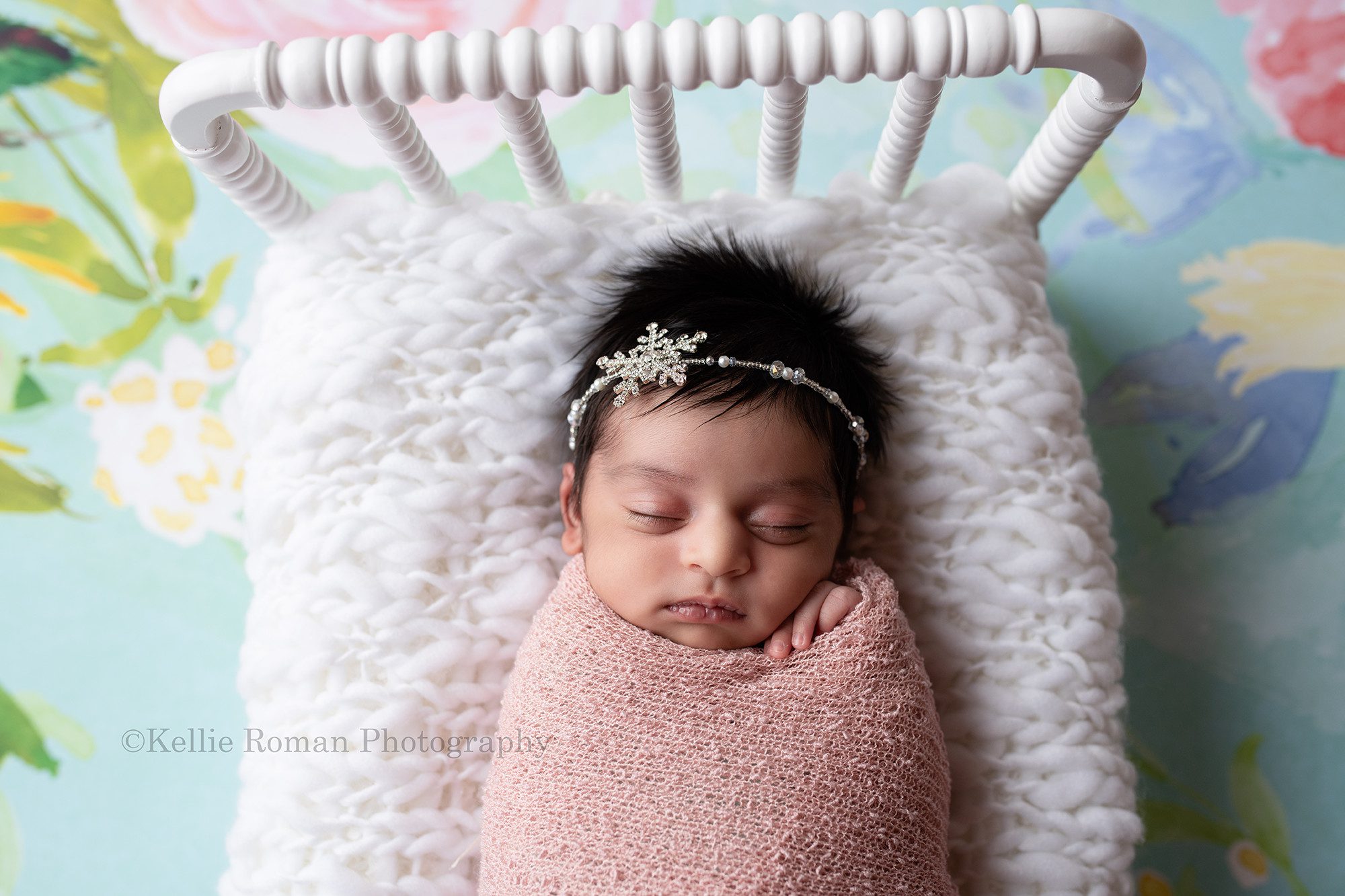 all wrapped up a newborn baby girl is in a milwaukee photographers studio located in greendale Wisconsin. The baby is wrapped in a pink swaddle and is sleeping on top of a white bed with a white knit blanket she has one had sticking out of the wrap. she has dark skin and lots of dark hair and has a rhinestone snowflake headband on