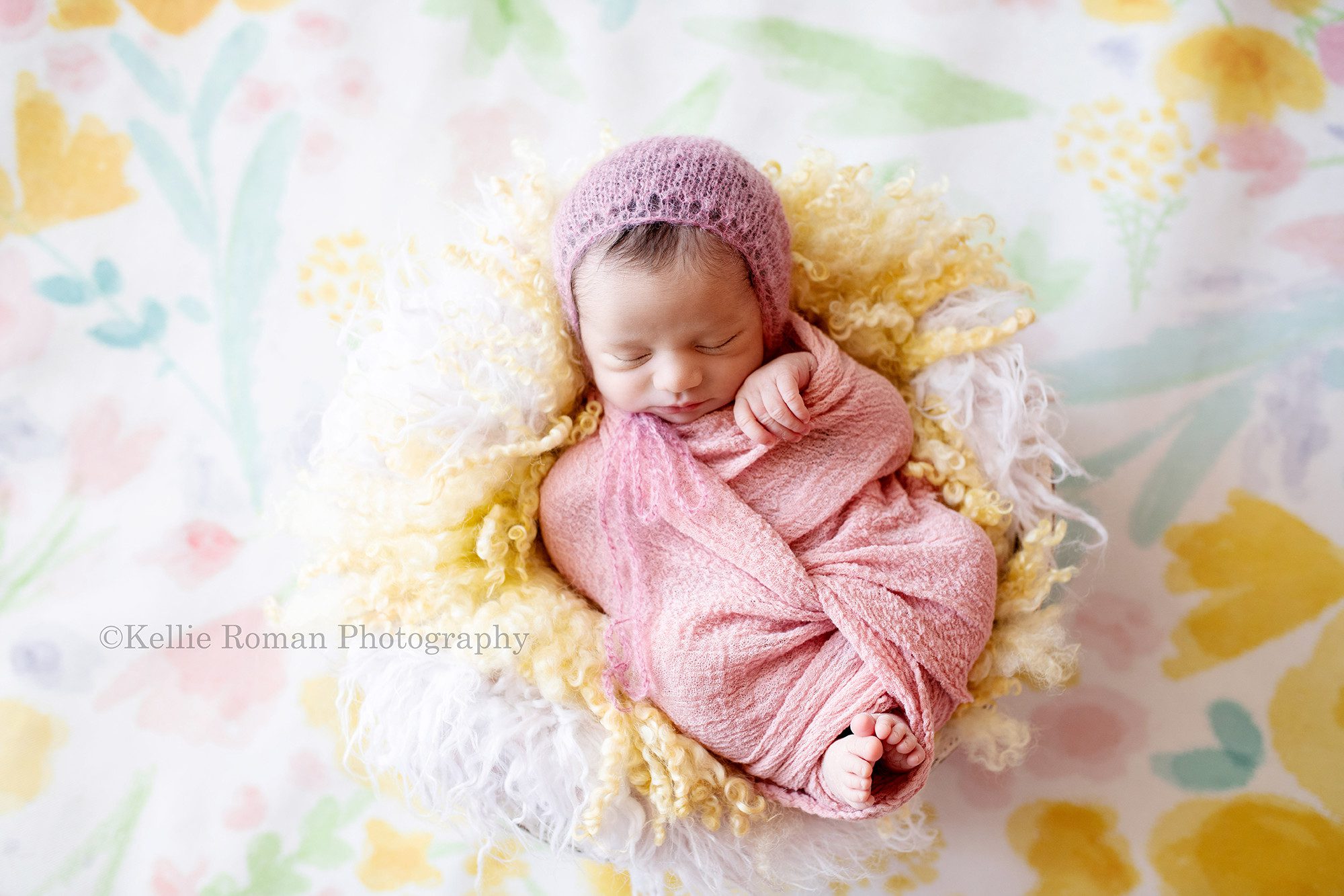 milwaukee newborn photographer. A baby girl is a milwaukee photographer studio In greendale is wrapped in a pink fabric with her toes sticking out of the bottom. She has a pink bonnet on, and is sleeping on her back in a tub with yellow and white fur. The tub is on a floor drop with pink green yellow and blue flowers on it.