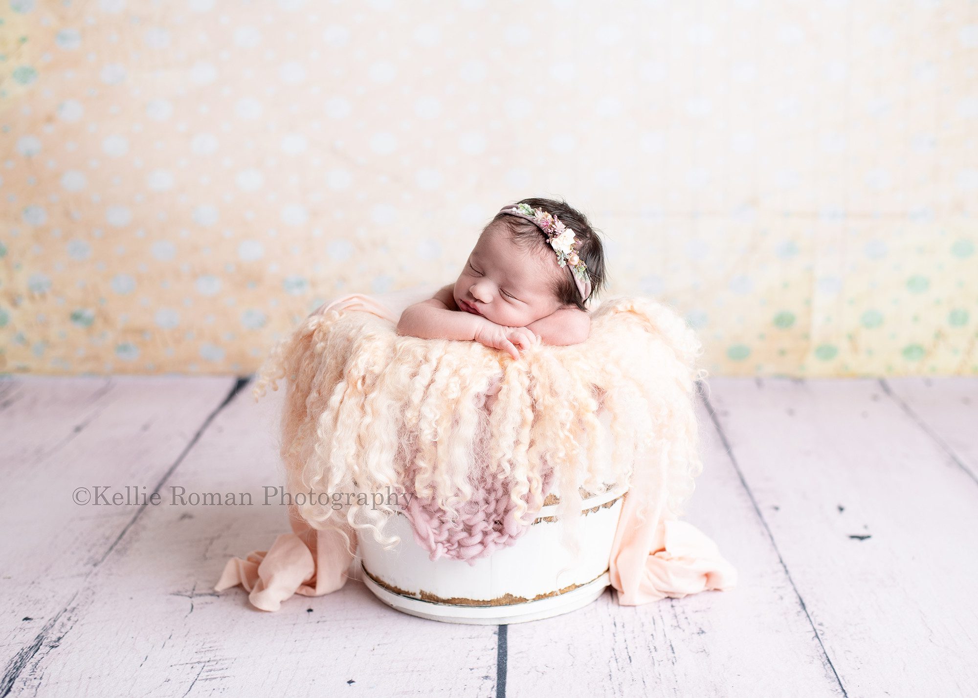 milwaukee newborn photographer. A infant baby girl is in a milwaukee photographer studio located in greendale. She's propped upright in a white bucket with peach and mauve fabric layers. The bucket is onto of a white rustic floor, and she's in front of a cream backdrop with teal polka dots.