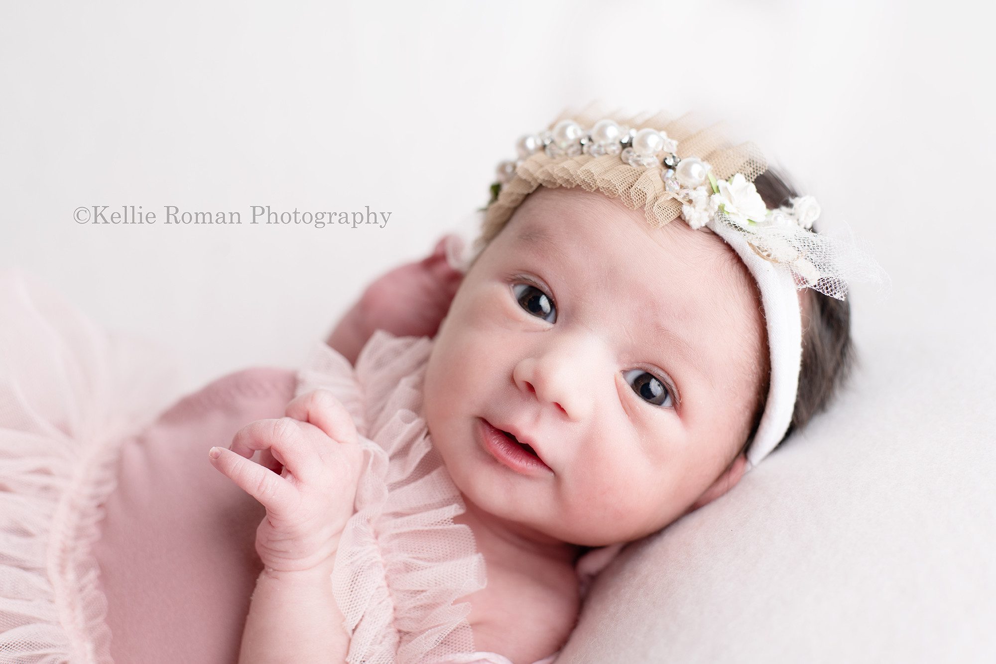 milwaukee newborn photographer. A baby girl in a milwaukee photographers studio located in greendale is laying on her back on a cream colored fabric while she's wide awake. She's wearing a mauve colored tutu romper, with a tule headband. She's looking into the camera with very good eye contact.