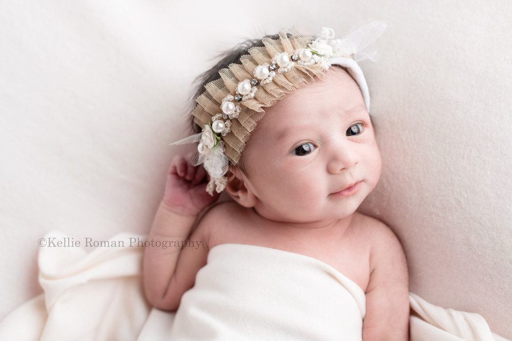 milwaukee newborn photographer. A infant baby girl is in a milwaukee photographer studio located in greendale Wisconsin. The baby is wide awake and laying on her back on top of cream fabric. She's wearing a cream colors lacy headband and is looking off to the side.