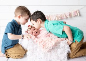 Milwaukee studio newborn photographer. A newborn baby girl and her two older brothers are in a milwaukee photographer studio in greendale Wisconsin. The baby girl is in a pink swaddle wrap and laying on her side in a white crate with white fur inside. The baby has a floral headband on. Her brothers have on a navy and teal polo shirts. The boys are both looking at her, one is leaning in and giving her a kiss. The backdrop is white wood with a pink and off white banner.