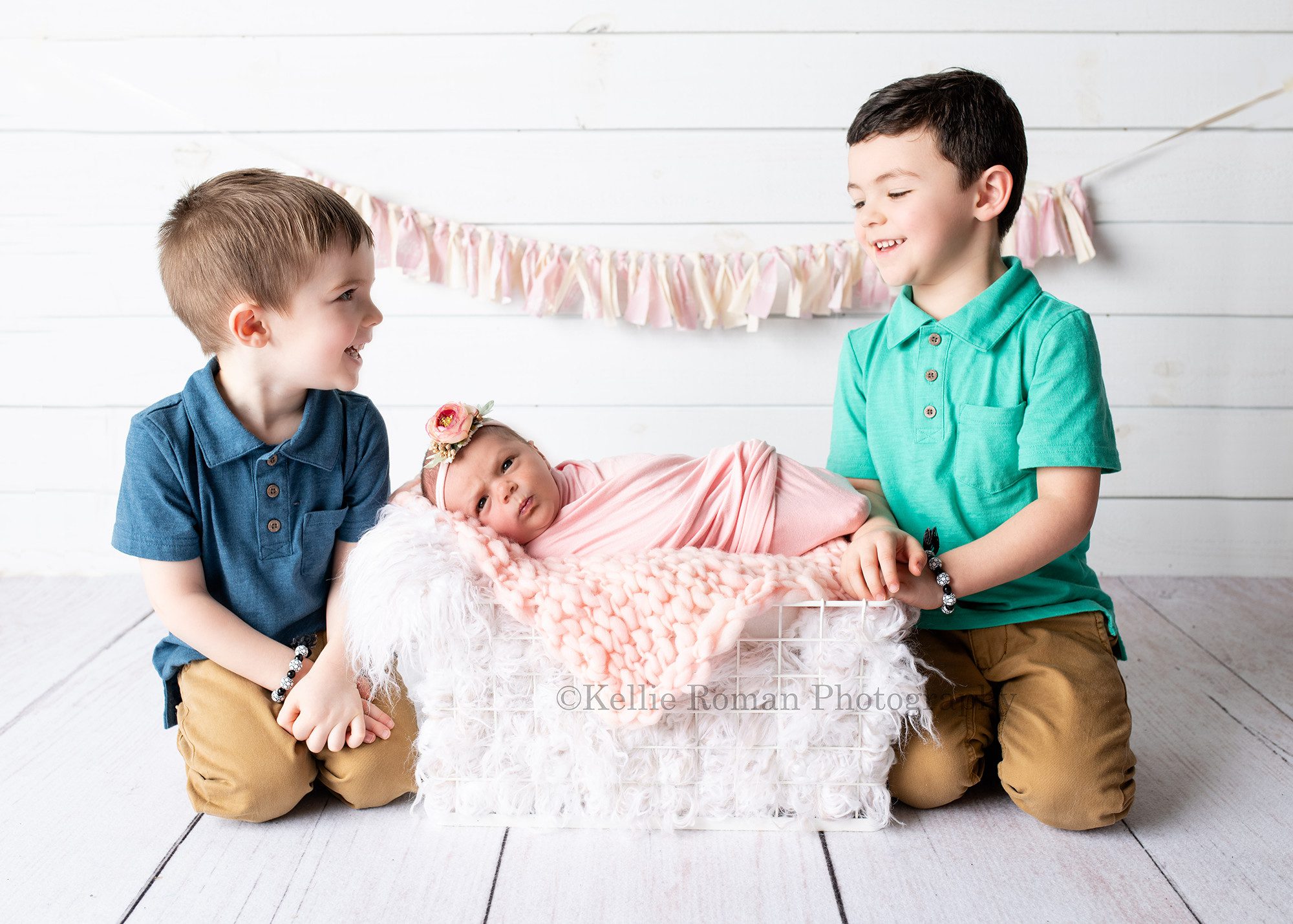 milwaukee studio newborn photographer. A newborn baby girl and her two toddler brothers are in a milwaukee photographer studio in greendale. The baby girl is wrapped in a light pink wrap, and has flower headband on. She is awake and laying on her side in a white wire crate filled with pink and white furs. Her brothers are on each side of the crate looking at each other smiling. The boys are wearing khakis and polo shirts that are blue and teal.