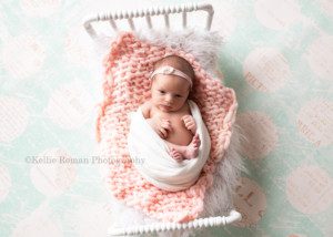 milwaukee studio newborn photographer. A baby girl is in a milwaukee photo studio located in greendale Wisconsin. The baby girl is wide awake laying on her back on a white little wood bed. There is white fur and light pink knitted blanket on top of the bed. The newborn is wrapped in a light pink fabric with her feet and hands sticking out. The bed is on top of a teal floor drop with big cream colored polka dots.