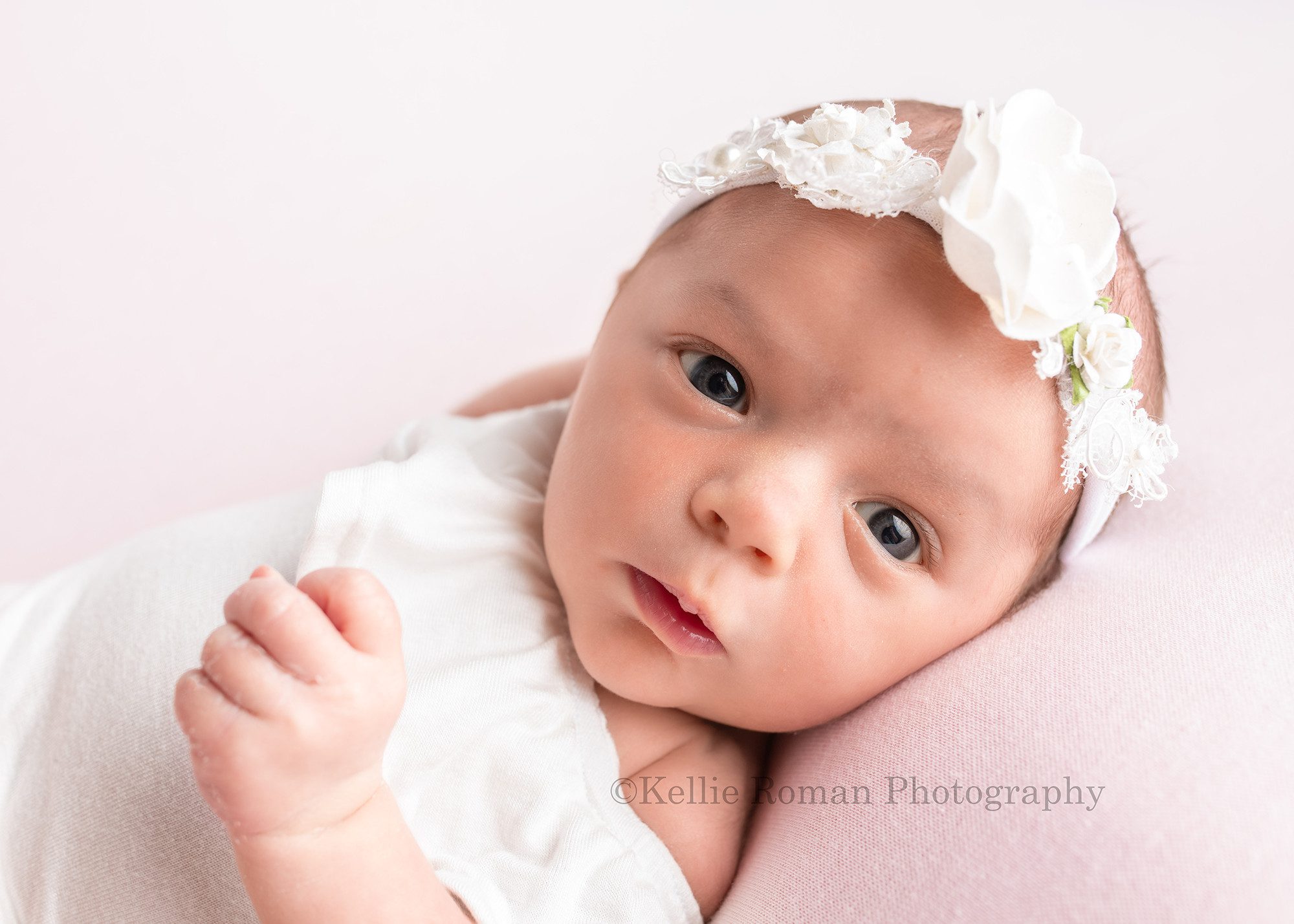 milwaukee studio newborn photographer. An infant baby girl is in a milwaukee photographers studio located in greendale Wisconsin. The baby girl is laying on her back with a white romper on and a white floral headband. she's on top of pink posing fabric, and is looking right into the lens of the camera