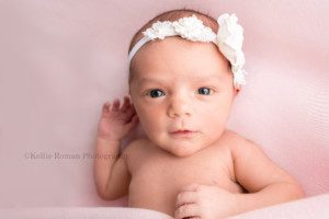 milwaukee studio newborn photographer. A newborn baby girl in a milwaukee photographer studio located in greendale Wisconsin. The baby girl is wide awake and laying on top of pink fabric. She's looking into the camera and has a white floral headband on.