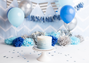uneasy cake smash the image is a cake smash set up for a one year old in a greendale Wisconsin photographers studio. it has shades of blues grey and silver. There is a chevron backdrop with banners and blue balloons. Blue tissue paper pom poms and silver confetti. A smash cake is on a white cake stand