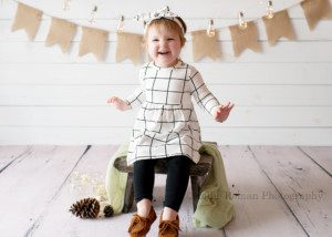 a 2 year old girl in a milwaukee photographer studio sitting on a white wood bench with a light green fabric on it. The girl has black leggings, with a white and black shirt and borrow moccasins. She is smiling really big and there is a pink cone next to her on the floor with a burlap banner and hanging string lights behind her