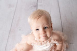 spring cake smash. a one year old girl is in a milwaukee photographers studio. The shot is close up from above the girl. she's wearing a light pink fluffy dress and sitting on a white wood floor. she's sucking in her bottom lip and has big blue eyes.