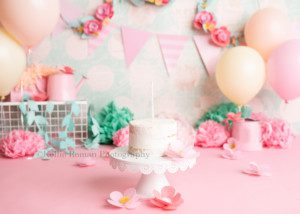 spring cake smash. The shot is a set up for a girls cake smash session. There is floral pink and teal decor around her with watering cans. The cake style is white frosted cake onto of a white cake stand.