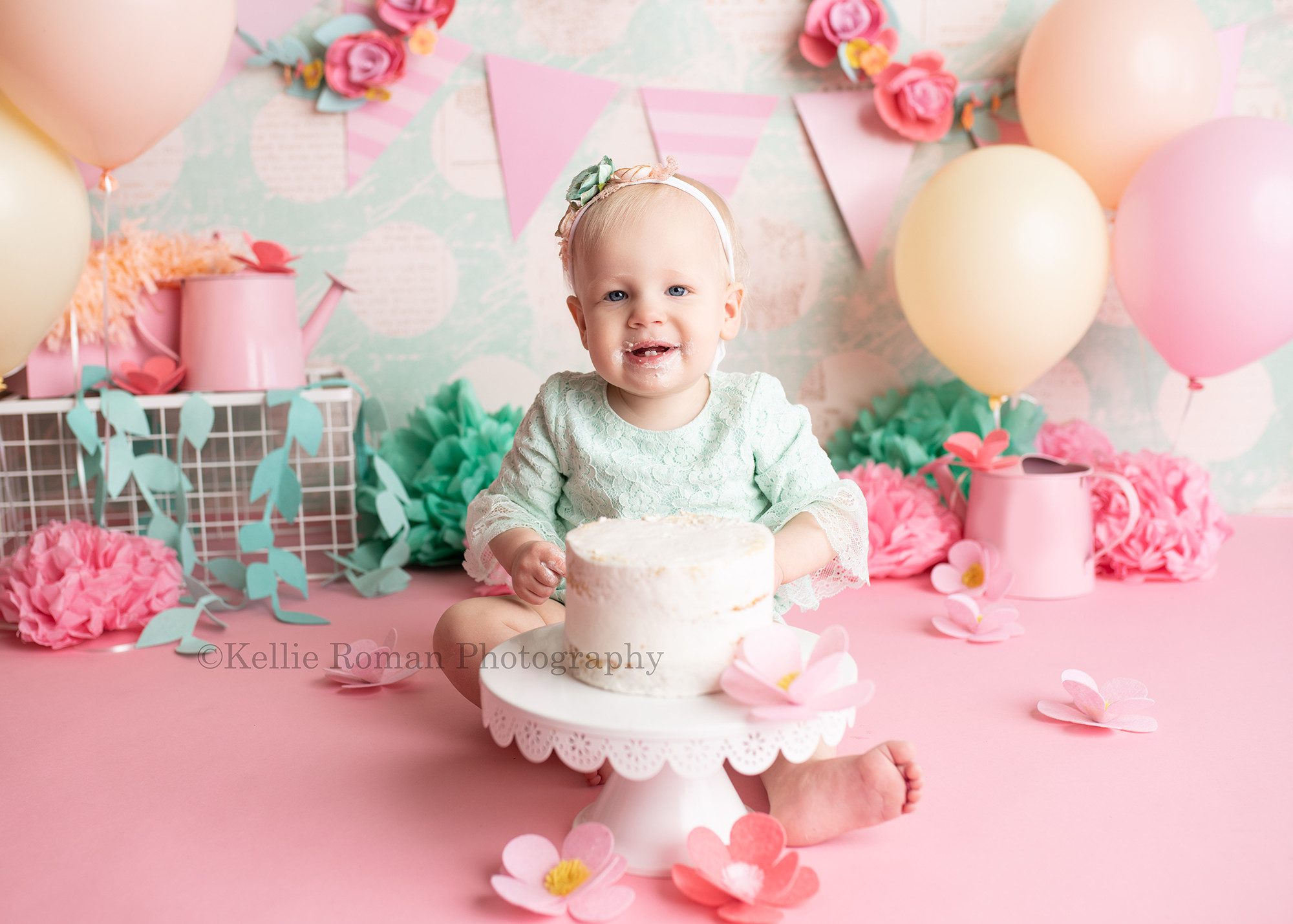 Spring cake smash. a one year old little girl is in a milwaukee photography studio having her cake smash photos taken. She's sitting in front of a teal polka dot backdrop with a pink banner and floral metal hoops. There are pastel peach, pink, and orange balloons. There is floral pink and teal decor around her with watering cans. She wearing a light teal romper and is sitting behind a cake style white frosted cake onto of a white cake stand. She's smiling at the camera