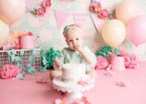 spring cake smash. a close up shot of a one year old smooshing cake into her face with her hand. she's in a milwaukee photographer studio for her milestone session. she's wearing a light mint lace romper and a matching headband. the backgrounders is bubblegum pink with teal and peach florals and tissue paper pom poms and pastel balloons. she is looking at the camera, while sitting behind a white frosted cake that is on top of a white cake stand.