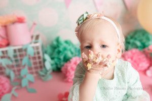 spring cake smash. a close up shot of a one year old smooshing cake into her mouth with her hand. she's in a milwaukee photographer studio for her milestone session. she's wearing a light mint lace romper and a matching headband. the backdrop and floor is bubblegum pink. There are teal pink and yellow flowers all over and pastel colored balloons.