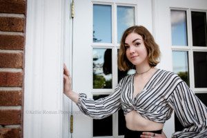 milwaukee senior photographers. a teenage girl is standing in front of a white window pain with her hand up on the frame. she's wearing a white top with black stripes and is showing off her midriff