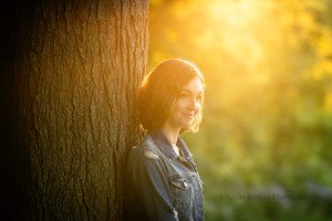 milwaukee senior photographers a teenage girl is in a Milwaukee County park standing and leaning against a tree. golden sunlight is coming from behind her, and she's looking over at her mom. she has short hair and jean jacket on