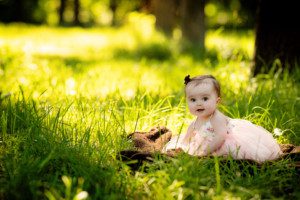 milwaukee family photographer. a 9 month old baby girl is sitting in tall grass in a park. she's wearing a light pink floral dress and has dark hair and eyes. she's smiling and looking at the camera