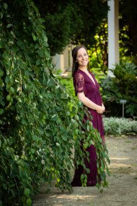 milwaukee family photographer. a women is wearing a long lace deep purple gown. she is smiling and holding her belly to show off her baby bump. she's standing by a dark green tree